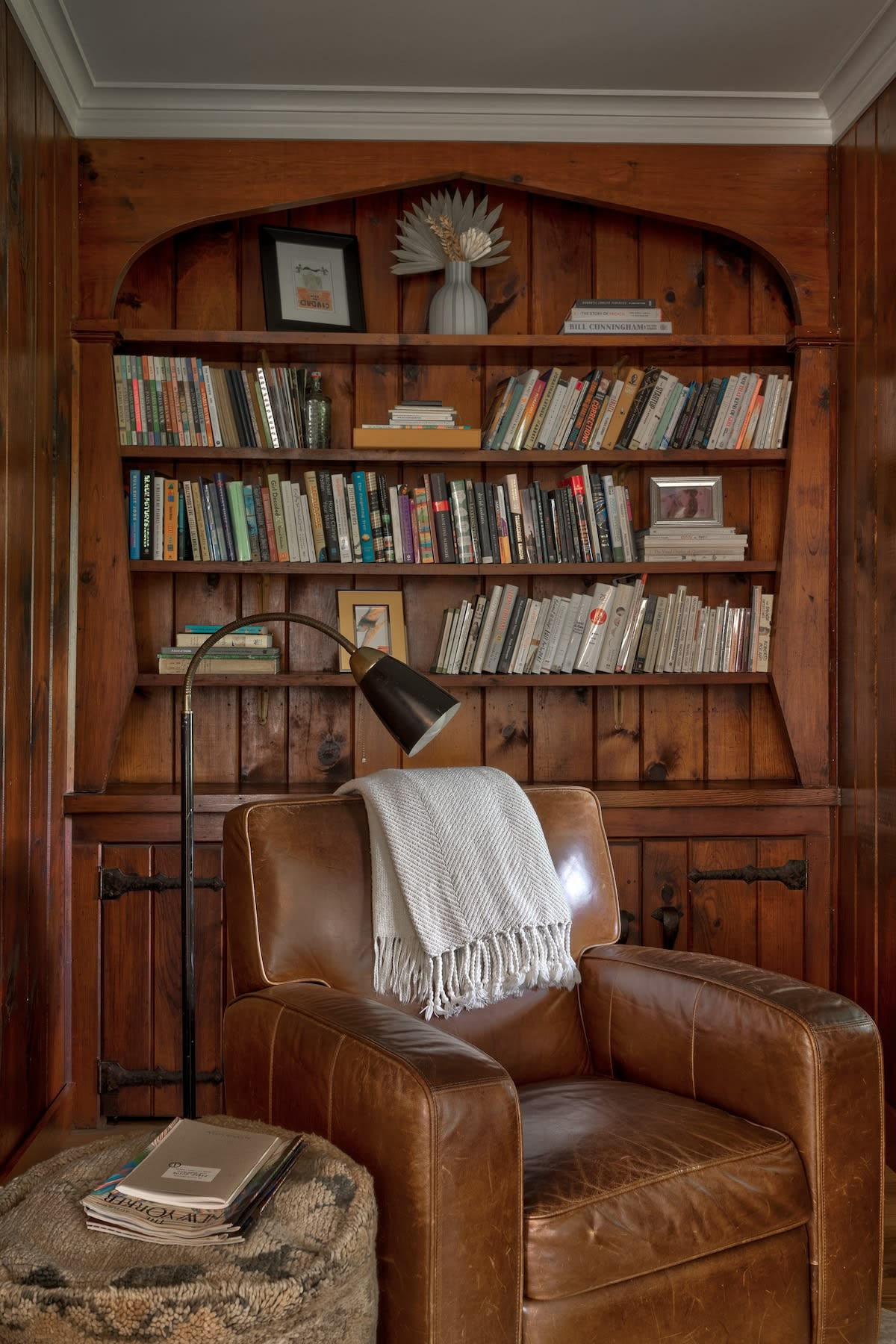 Inviting reading nook featuring a comfortable brown leather armchair, a floor lamp, and a built-in wooden bookshelf filled with books. The room is characterized by warm wood paneling, creating a cozy atmosphere.