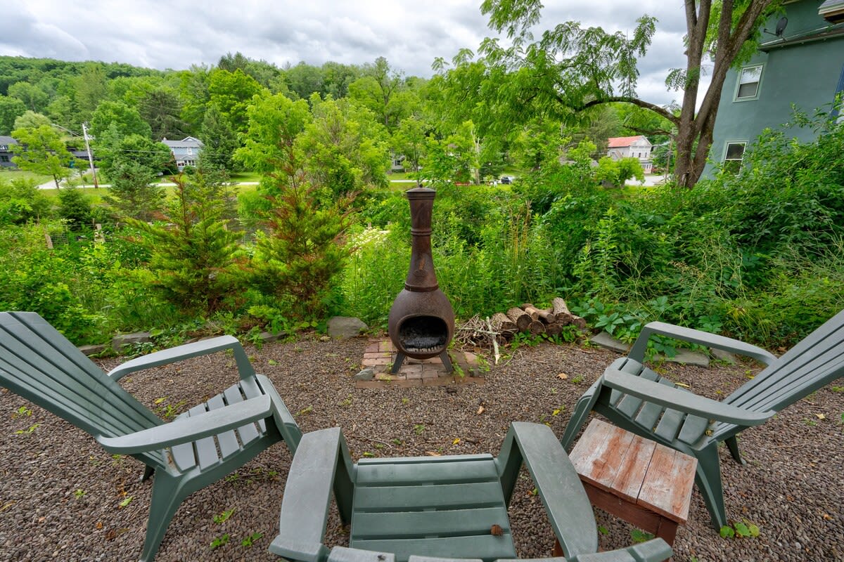 An outdoor seating area with three gray Adirondack chairs facing a tall, rustic kiva-style fire pit on a gravel surface, surrounded by lush greenery and distant views of a lake.