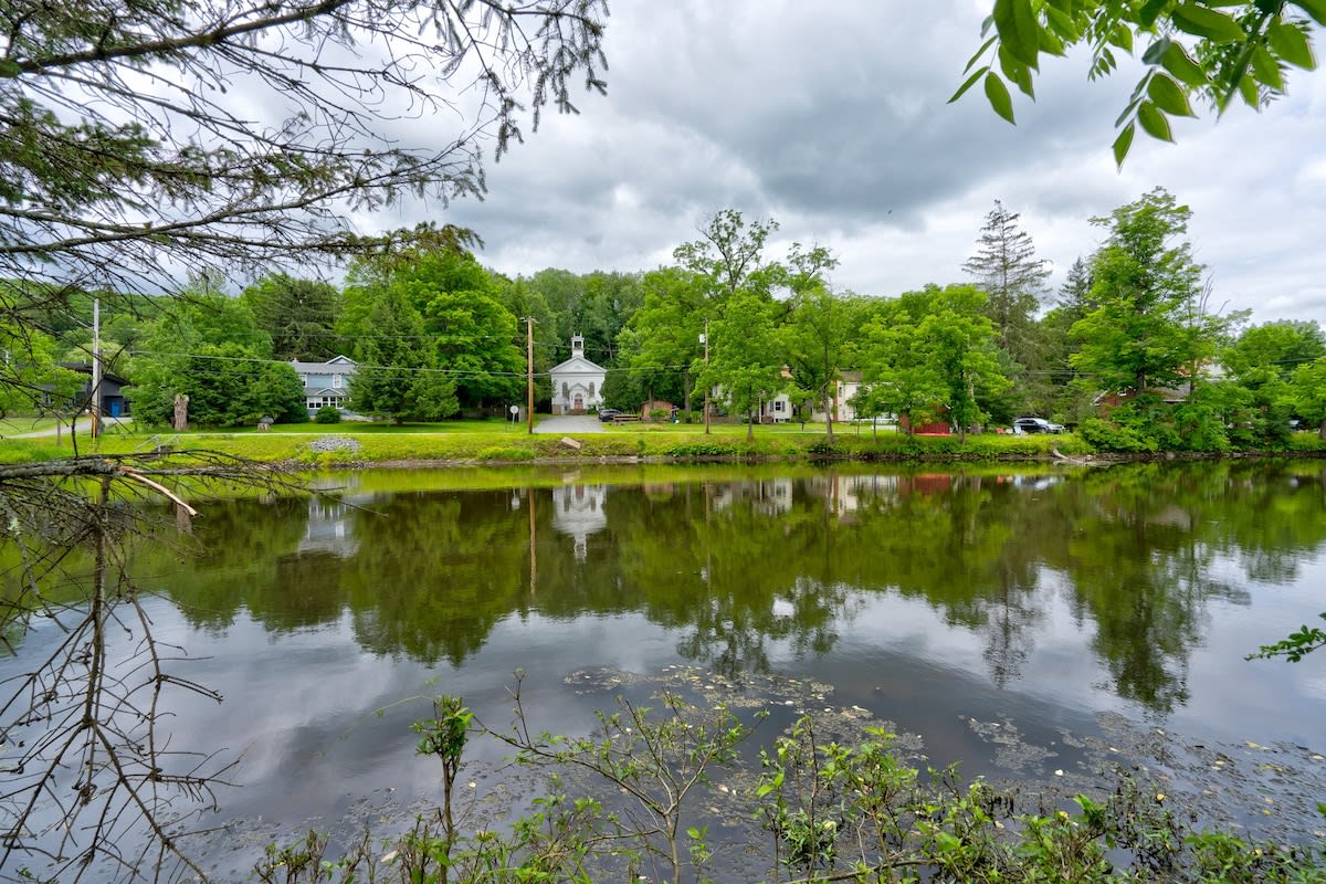 A scenic view across a calm lake reflecting the cloudy sky, towards a charming village with a white church and houses nestled among lush green trees. The foreground features natural foliage and branches.