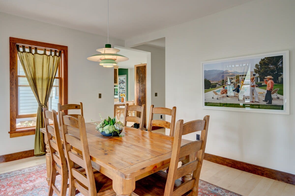 Spacious dining room featuring a rustic wooden dining table surrounded by six matching chairs, set atop a colorful patterned area rug. A distinctive modern pendant light illuminates the space, with two windows framed by yellow curtains.