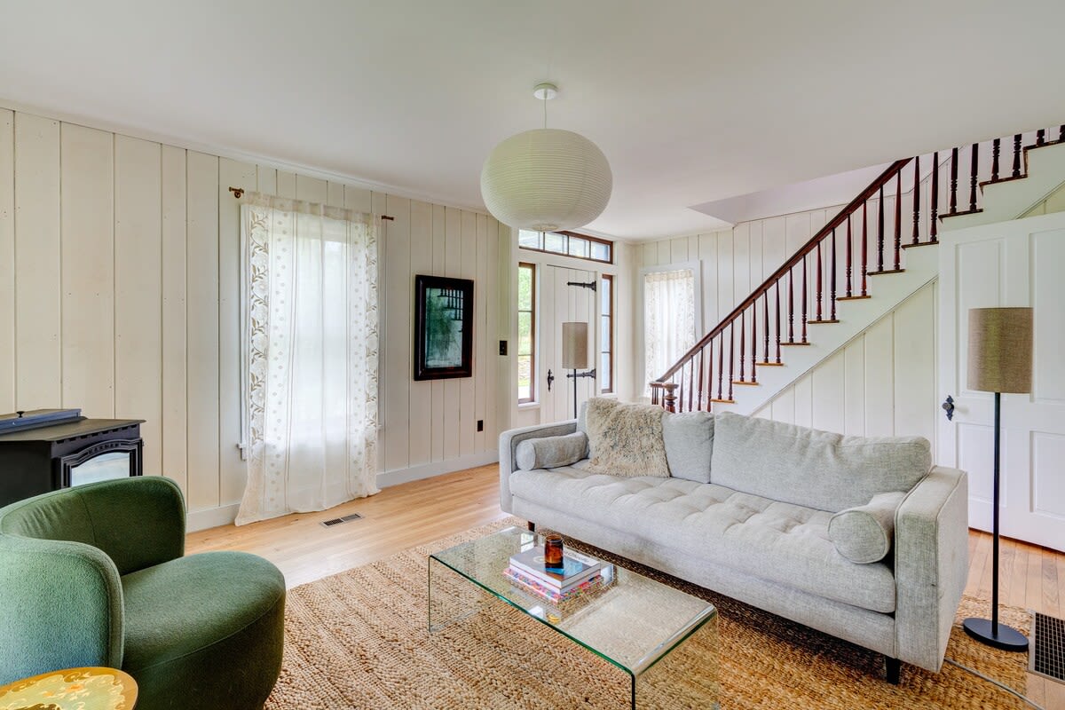 Inviting living room with a light grey sofa, a green armchair, and a modern glass coffee table on a natural jute rug. A prominent paper lantern light fixture hangs from the ceiling, and a wooden staircase is visible in the background.