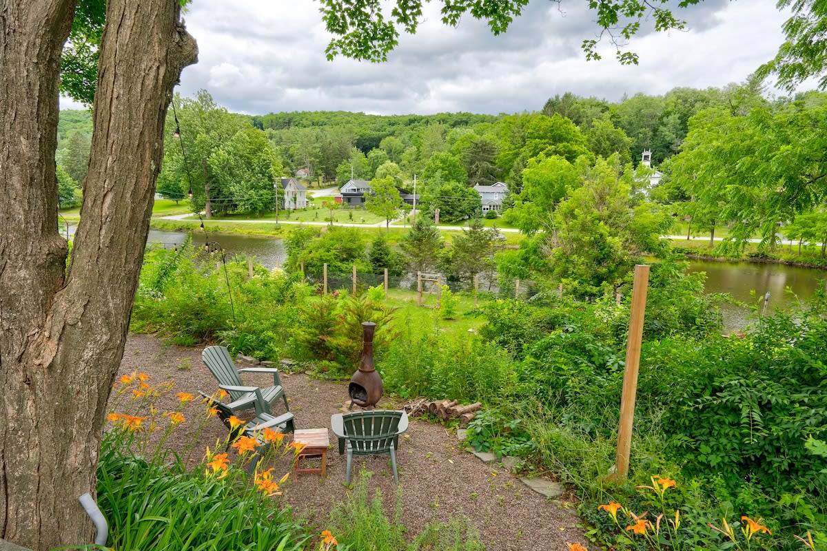 A gravel patio area features two green Adirondack chairs and a rustic metal fire pit, surrounded by lush green foliage and orange daylilies, offering a scenic view of a tranquil lake and distant houses under a cloudy sky.
