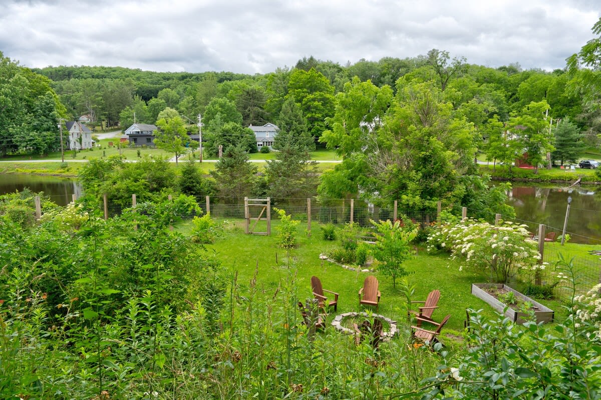 An expansive backyard garden area with lush green foliage, a fenced-in section, and a fire pit surrounded by Adirondack chairs. A lake is visible in the background, bordered by dense trees and distant houses.