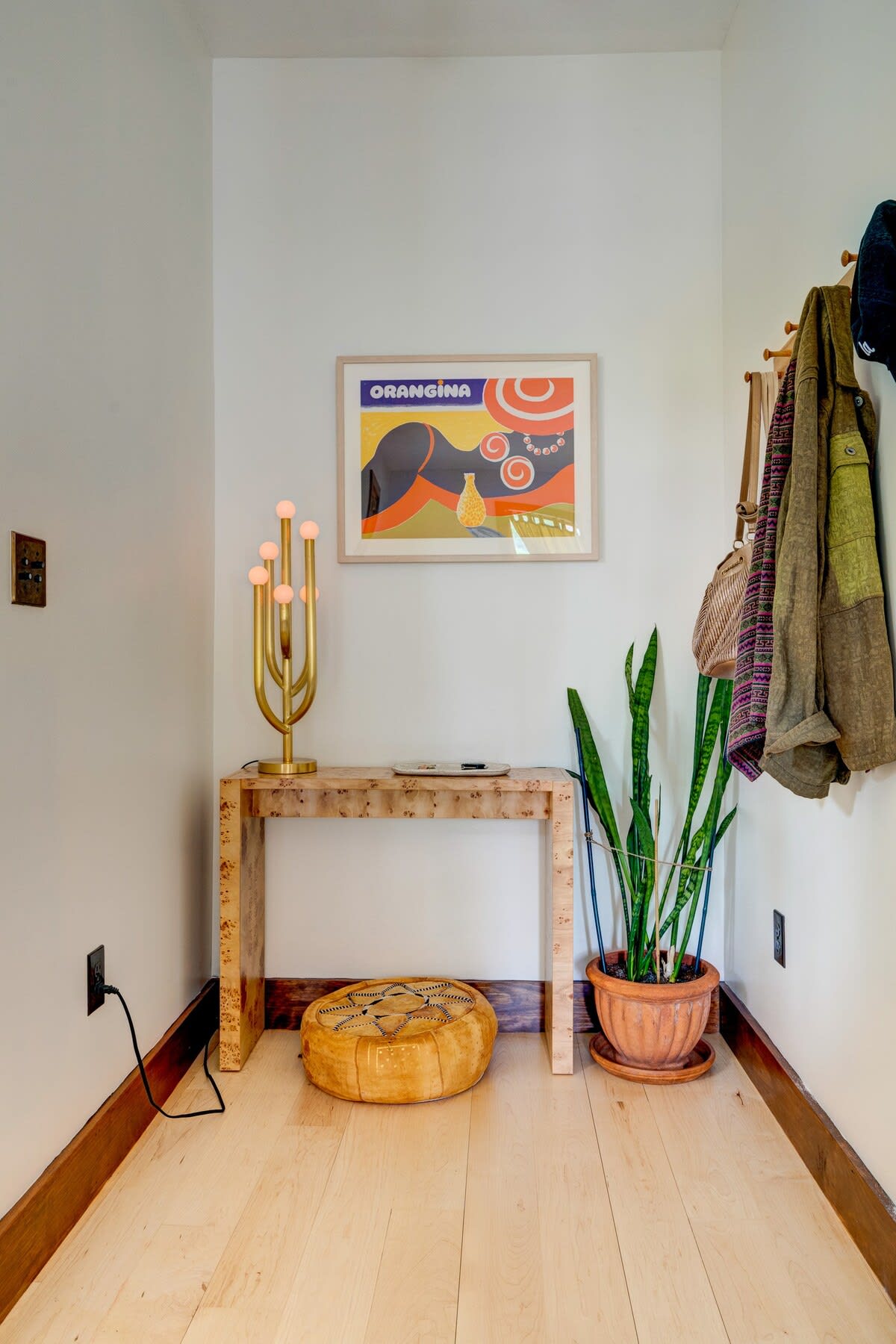 A welcoming entryway with light hardwood floors and white walls, featuring a rustic wooden console table with a modern gold lamp and a potted snake plant. A colorful "Orangina" art print hangs above the table.