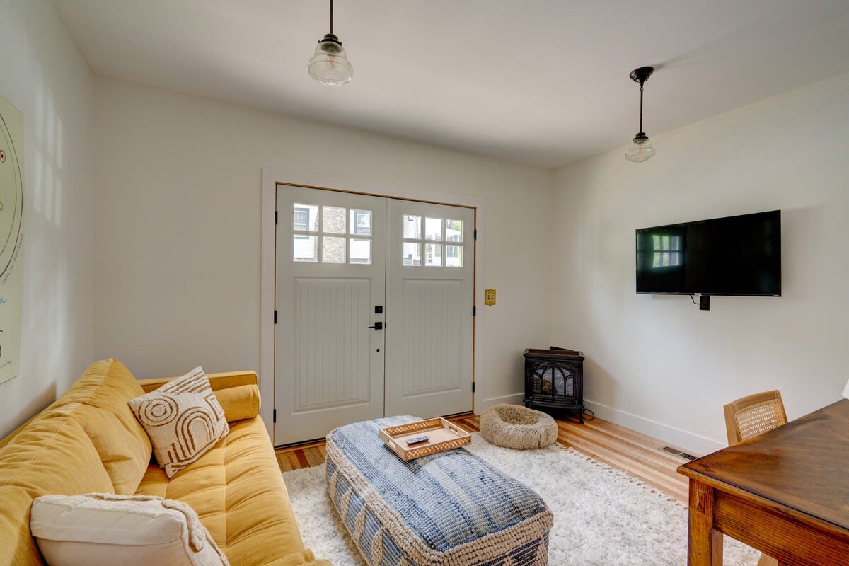 Cozy living space featuring a vibrant yellow sofa, a patterned woven ottoman, and a wall-mounted television. Double doors with glass panes lead outside, and light wood floors are covered by a shaggy rug.