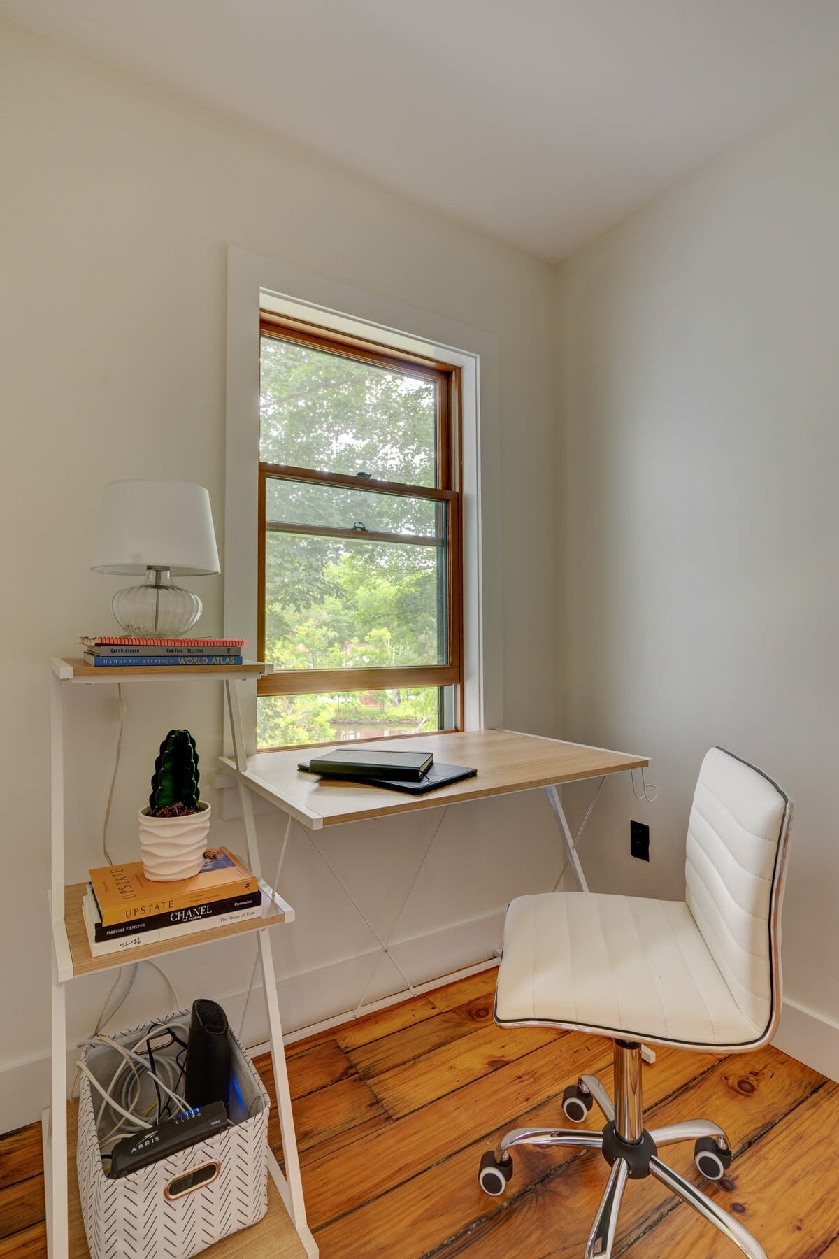 A bright home office space with light hardwood floors and white walls, featuring a minimalist wooden desk positioned in front of a window, and a white ergonomic office chair. A small white shelf holds books and a cactus.