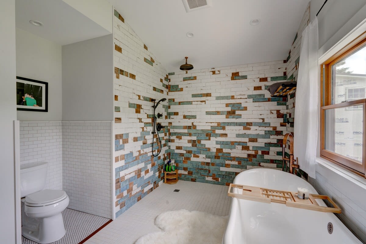 A modern bathroom featuring a freestanding white bathtub and a walk-in shower with unique white, brown, and blue patterned subway tiles. A toilet is visible in the foreground with white subway tile wainscoting.