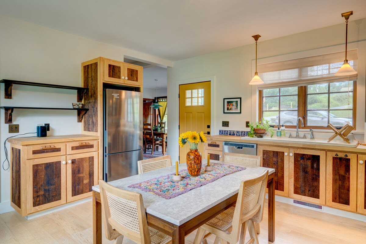 Expansive kitchen showcasing a stainless steel refrigerator, custom wood cabinetry with open shelving, and a dining table with chairs. A bright yellow door adds a pop of color to the space.
