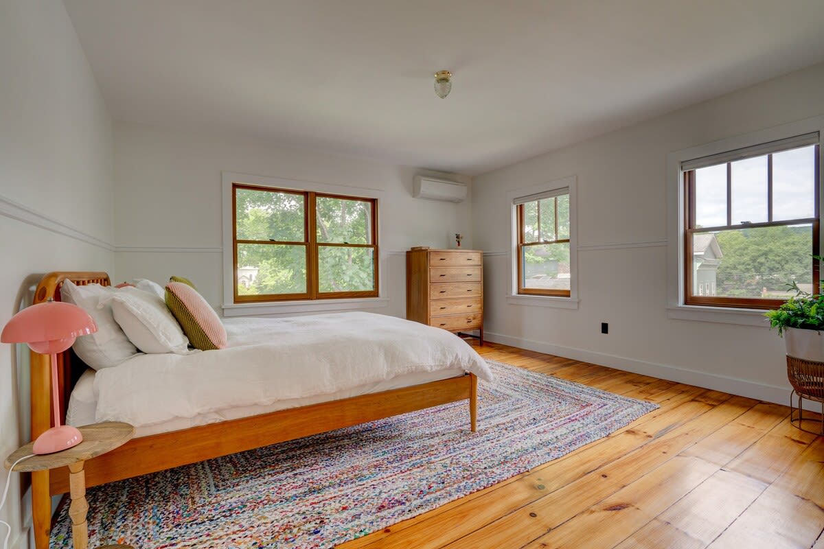 A wider view of the bright bedroom, showcasing the wooden bed with white bedding and a colorful area rug on light hardwood floors. Multiple windows provide ample natural light and views of the surrounding trees.