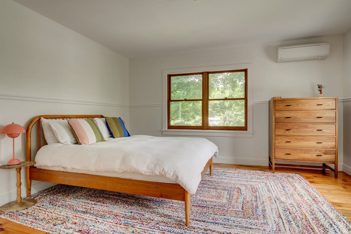 A bright bedroom with light hardwood floors and white walls, featuring a wooden bed frame with white bedding and colorful accent pillows. A tall wooden dresser stands against the far wall, and a window offers views of greenery.