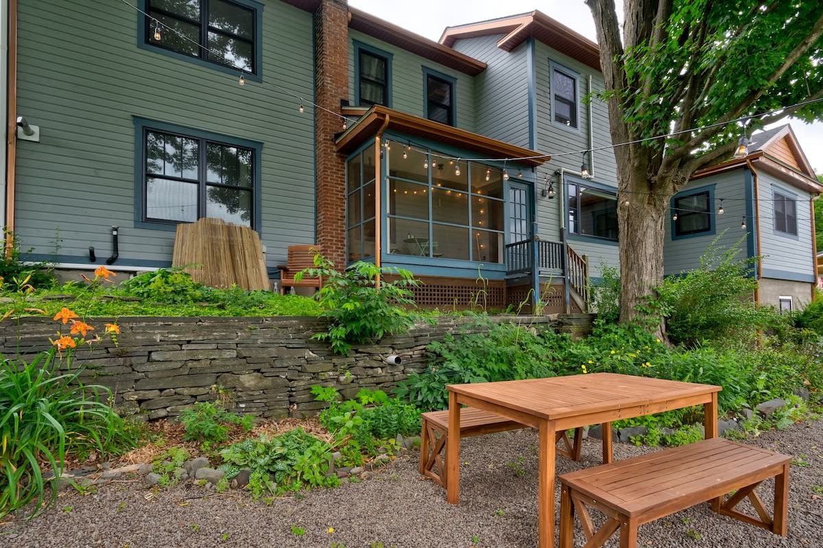 The back of a light blue house with dark trim, featuring a screened-in porch with string lights, a stone retaining wall, and a gravel patio with a wooden picnic table and benches, surrounded by lush greenery.