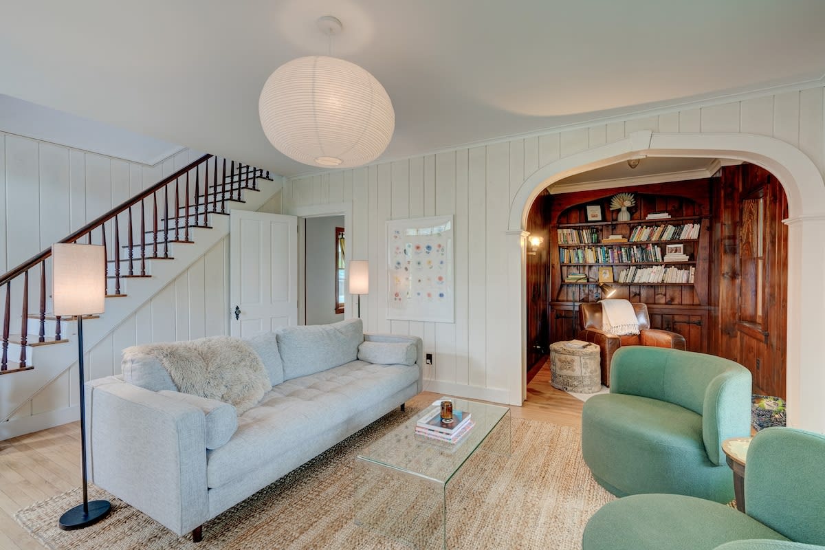 A bright living room with white vertical paneling, a light gray sofa, a glass coffee table, and a large paper lantern light, with a staircase leading upstairs and an arched doorway to a wood-paneled room.