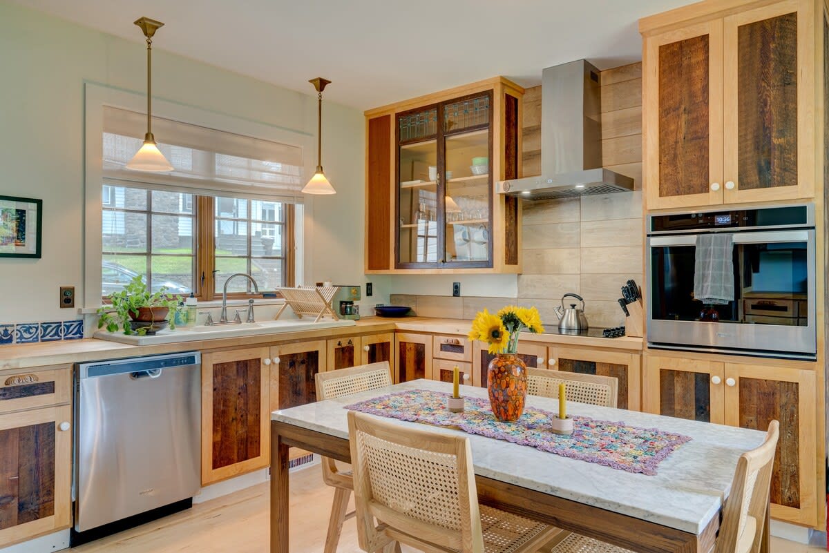 Bright kitchen featuring custom light and dark wood cabinetry, stainless steel appliances including a dishwasher and built-in oven, and a large window above the sink. A dining table with four chairs is centered in the room.