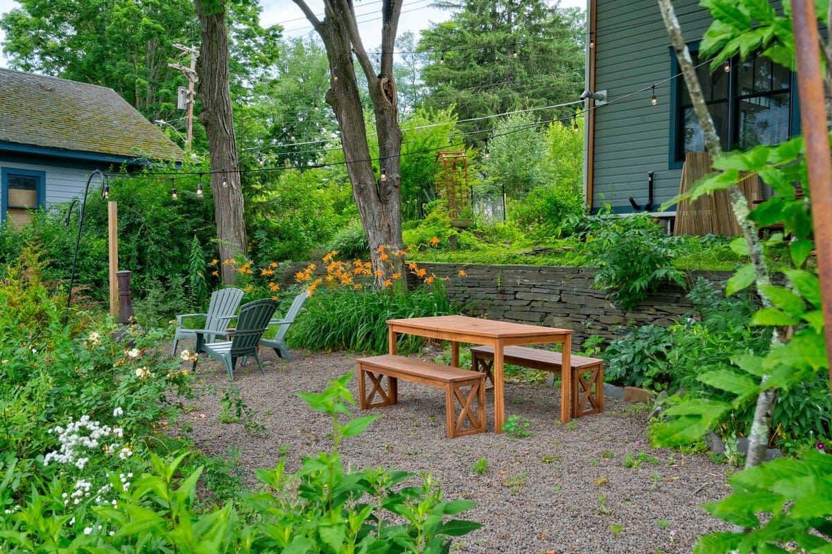 A spacious gravel patio features a wooden picnic table with two matching benches, nestled among vibrant green plants and trees, with string lights overhead and a portion of the house exterior visible in the background.