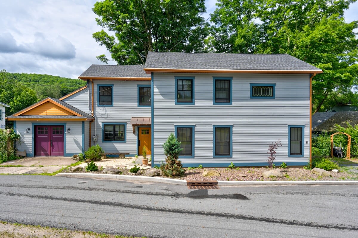 The front exterior of a light blue two-story house with dark blue trim and a vibrant purple garage door, situated on a paved street with a small driveway, surrounded by green trees and a cloudy sky.