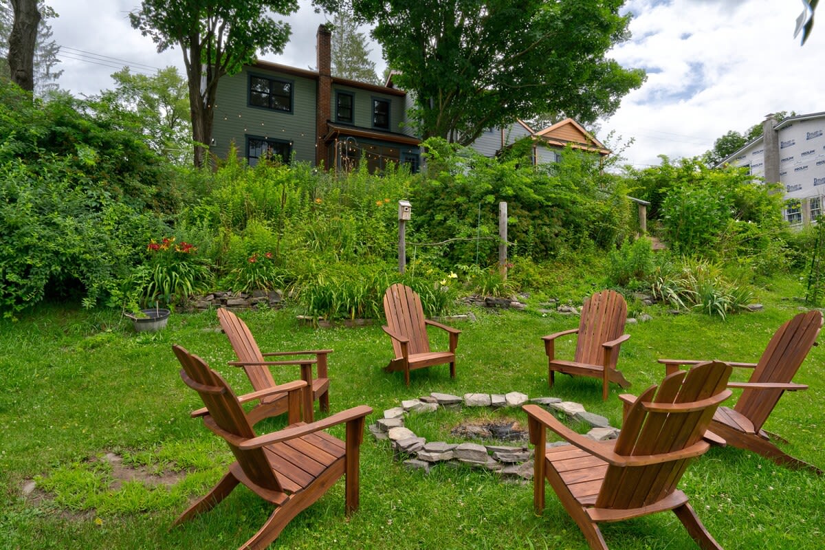 A circular arrangement of six wooden Adirondack chairs surrounds a stone-lined fire pit on a well-maintained grassy lawn, with a lush, tree-covered hillside and a multi-story house visible in the background.