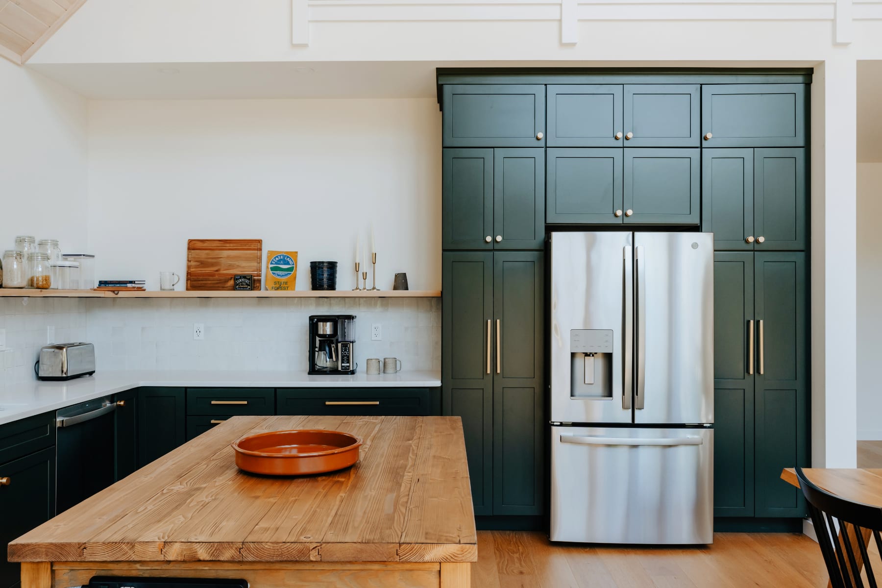 A modern kitchen features dark green shaker-style cabinetry, a large stainless steel French door refrigerator, and a rustic wooden island with a terracotta bowl.
