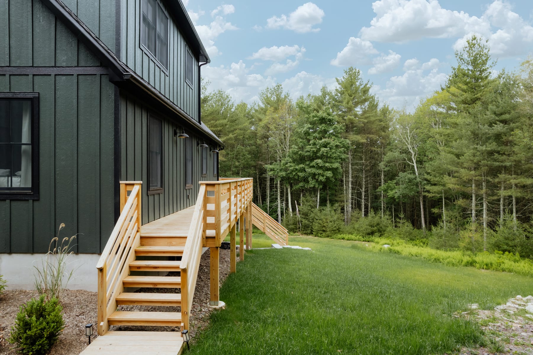 Side view of the dark green modern chalet, showcasing a long wooden deck with stairs leading down to a grassy yard and dense forest in the background.