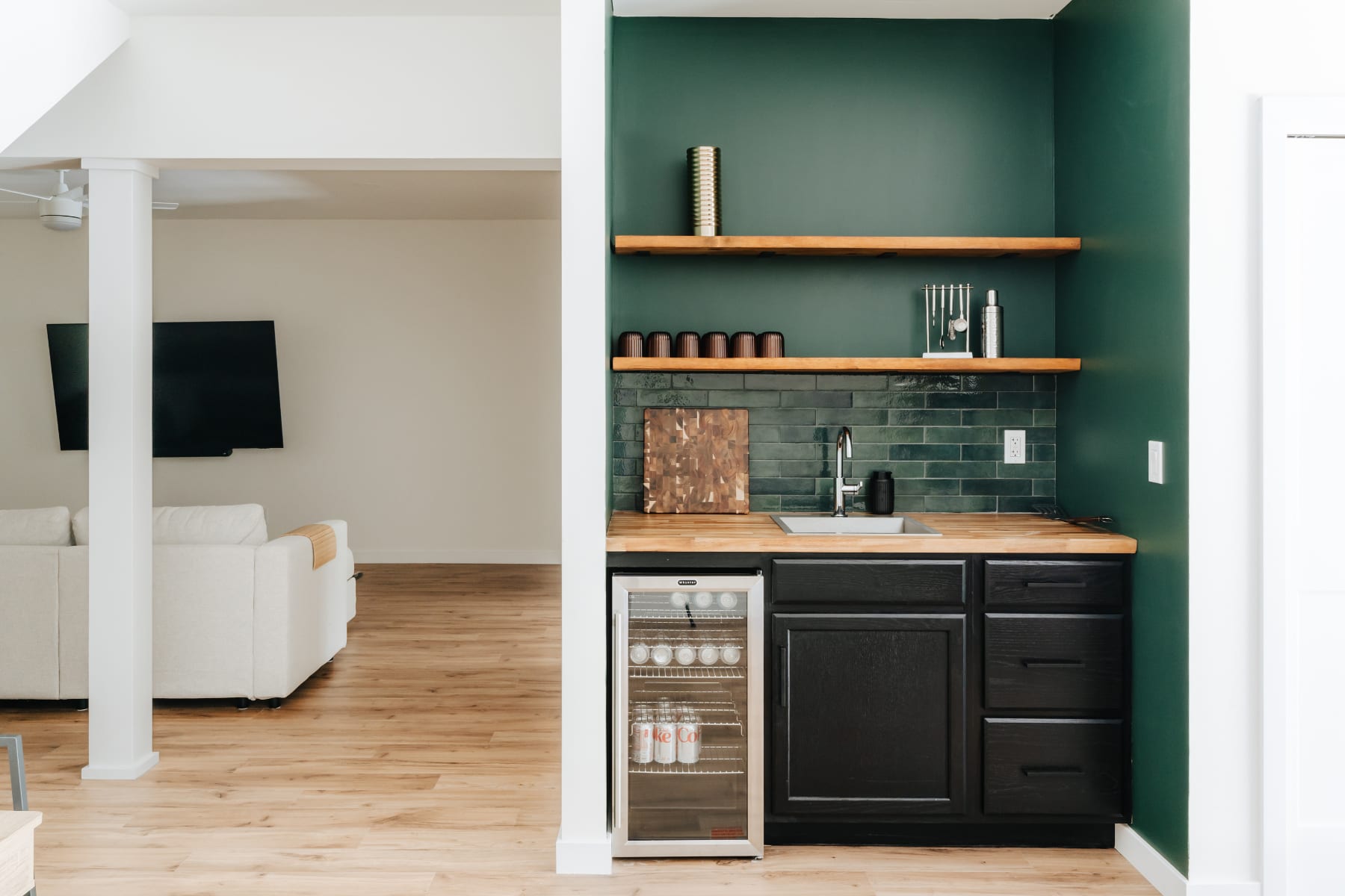A stylish wet bar area featuring a dark green accent wall, a wooden countertop with a sink, black base cabinets, open wooden shelving, and a glass-front mini-fridge, adjacent to a living space.