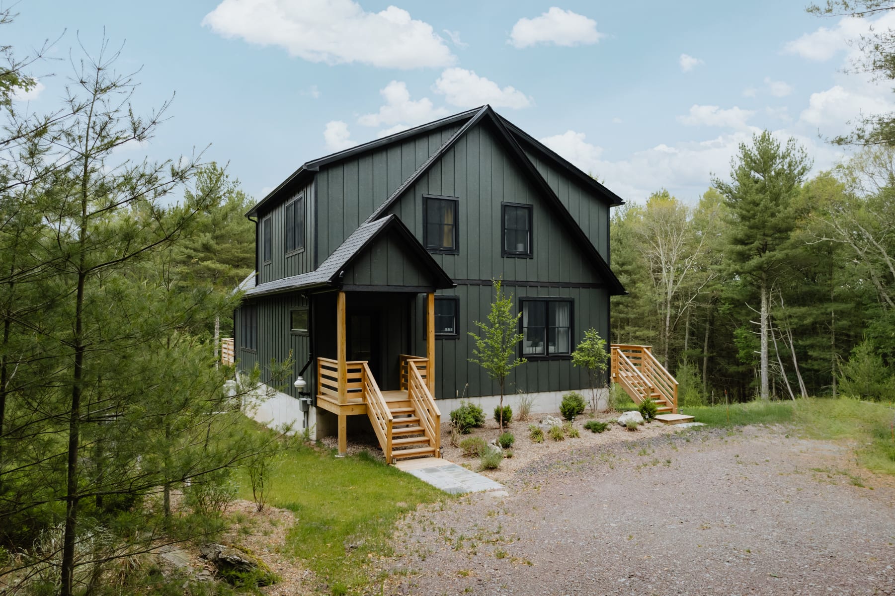 Angled front view of the modern dark green chalet, showcasing its striking A-frame architecture, black windows, and a covered front porch with wooden stairs, set amidst a natural, wooded environment.
