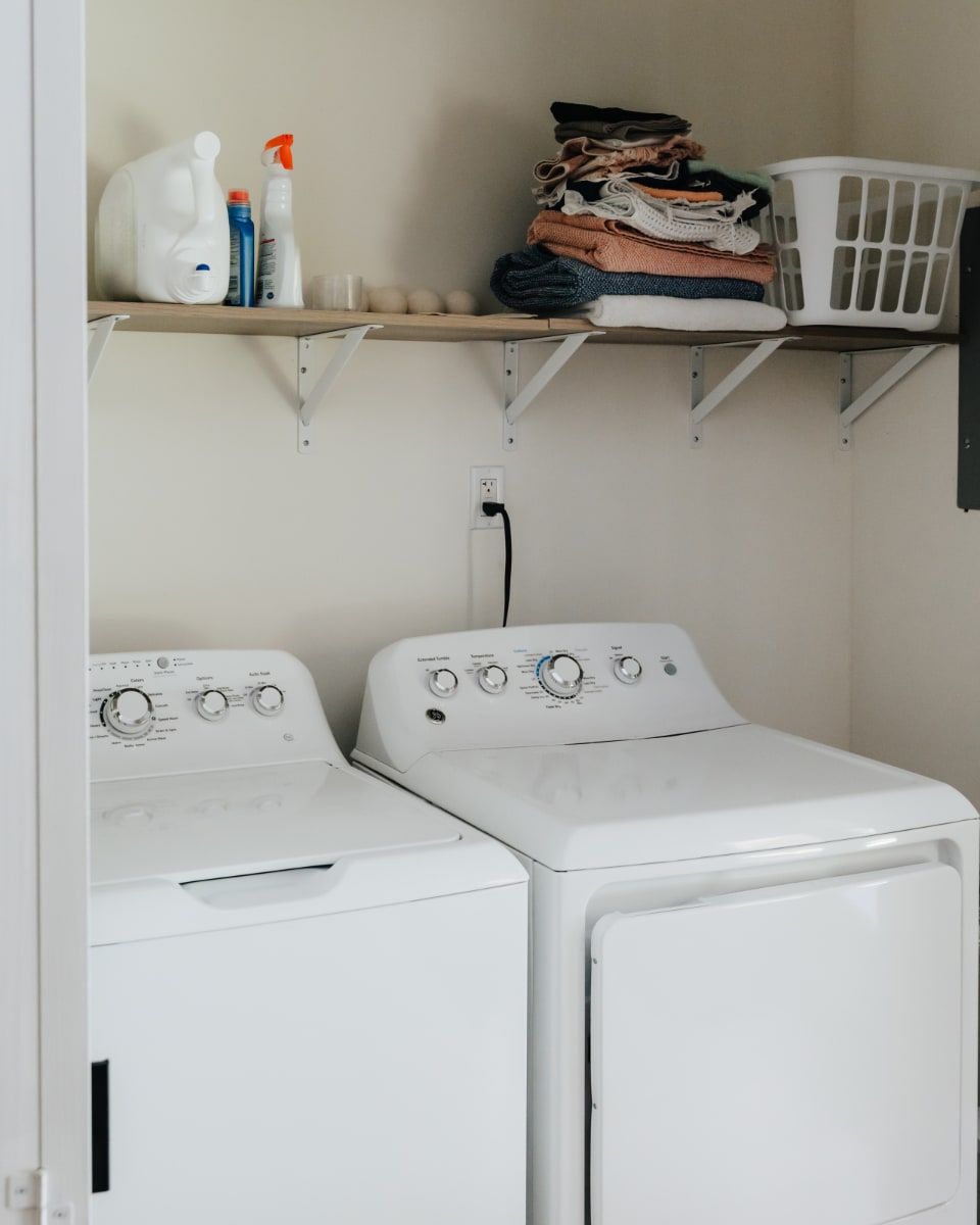 Functional laundry room featuring a white top-load washing machine and front-load dryer, with a wooden shelf above holding laundry supplies and folded clothes.