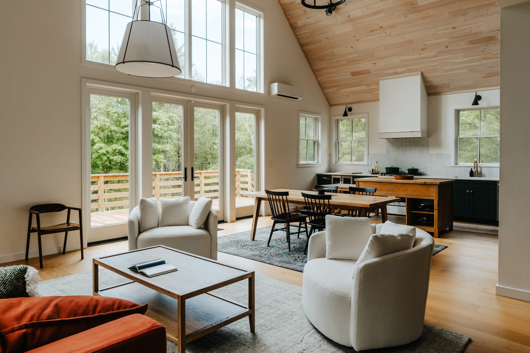 An expansive open-concept living area boasts a vaulted wood-paneled ceiling, large windows overlooking a lush forest, and comfortable white armchairs arranged around a light wood coffee table.