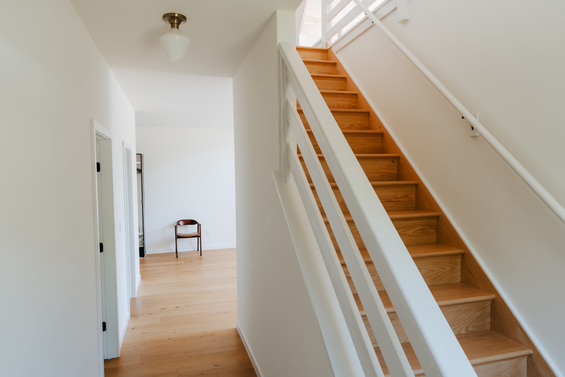 A bright hallway with light hardwood floors and white walls, leading to a wooden staircase with white railings, illuminated by a simple ceiling light fixture.