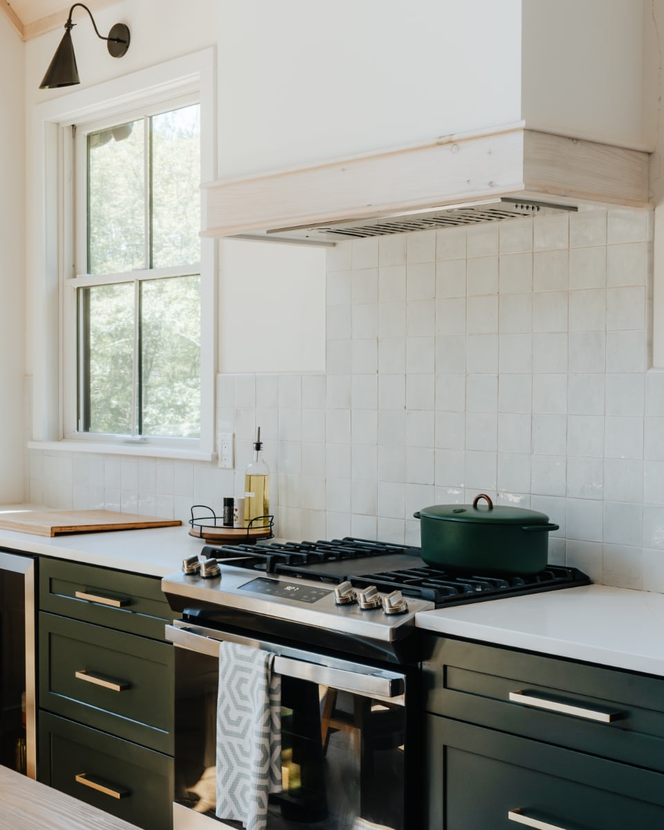 A stainless steel gas range is centered in the kitchen, flanked by dark green cabinets and white countertops, with a custom light wood range hood above and a window providing natural light.