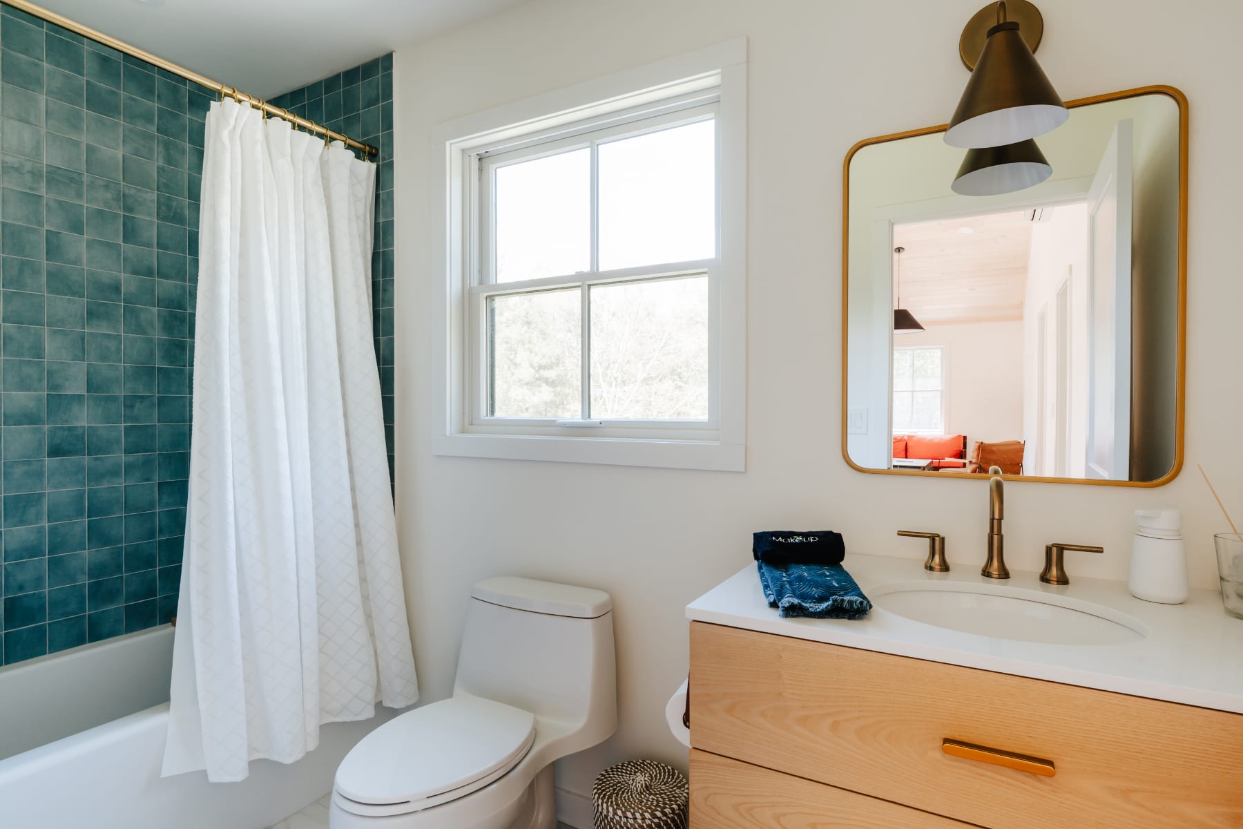 A modern bathroom featuring a white toilet, a light wood vanity with a white sink and gold faucet, a rectangular mirror with a brass light fixture, and a shower/tub combo with dark teal square tiles and a white curtain. A window provides natural light.
