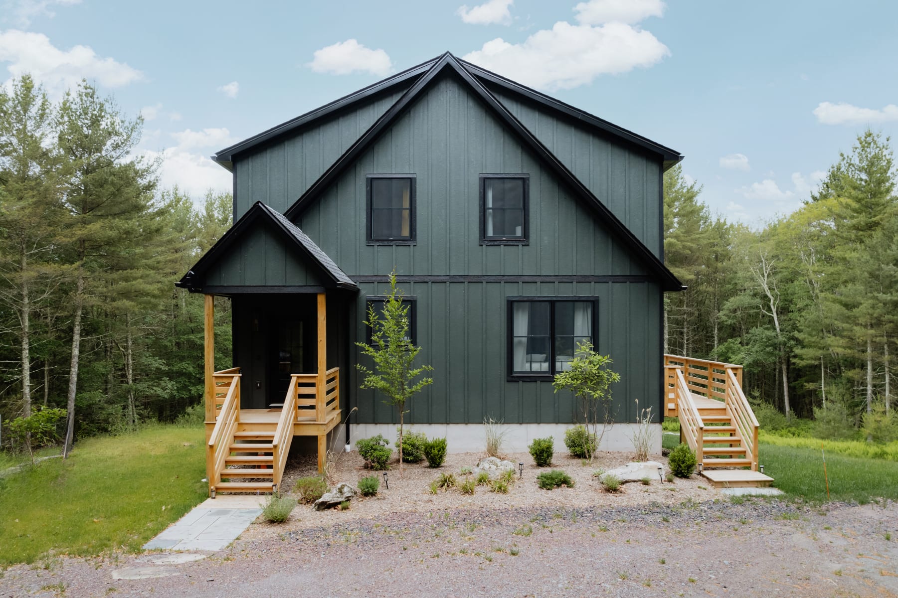 Front facade of the modern dark green chalet, featuring a prominent A-frame design, black-framed windows, and a covered entrance with wooden stairs, surrounded by minimal landscaping and a gravel driveway.