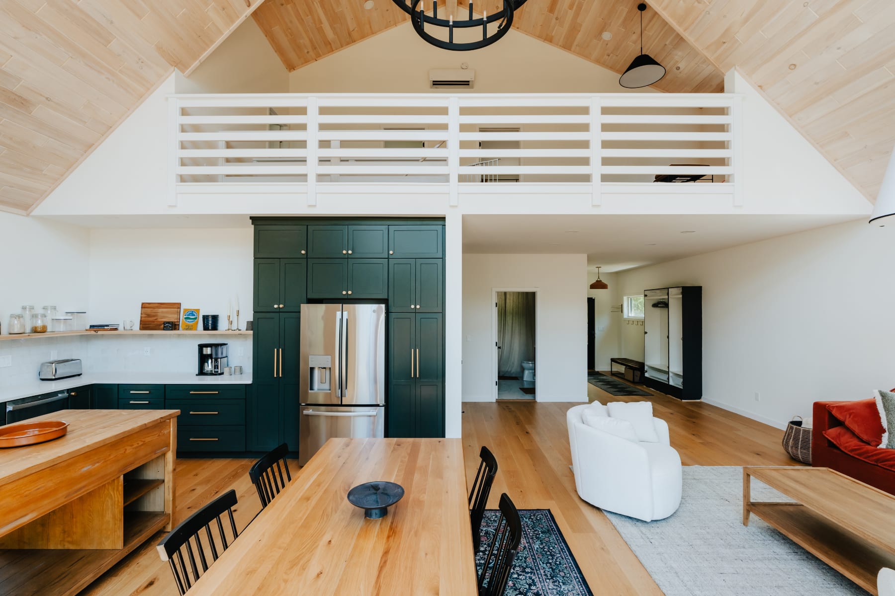 Expansive open-concept space with a vaulted wood ceiling, dark green kitchen cabinetry, a large dining table, and a living area, featuring a white railing loft overlooking the main floor.