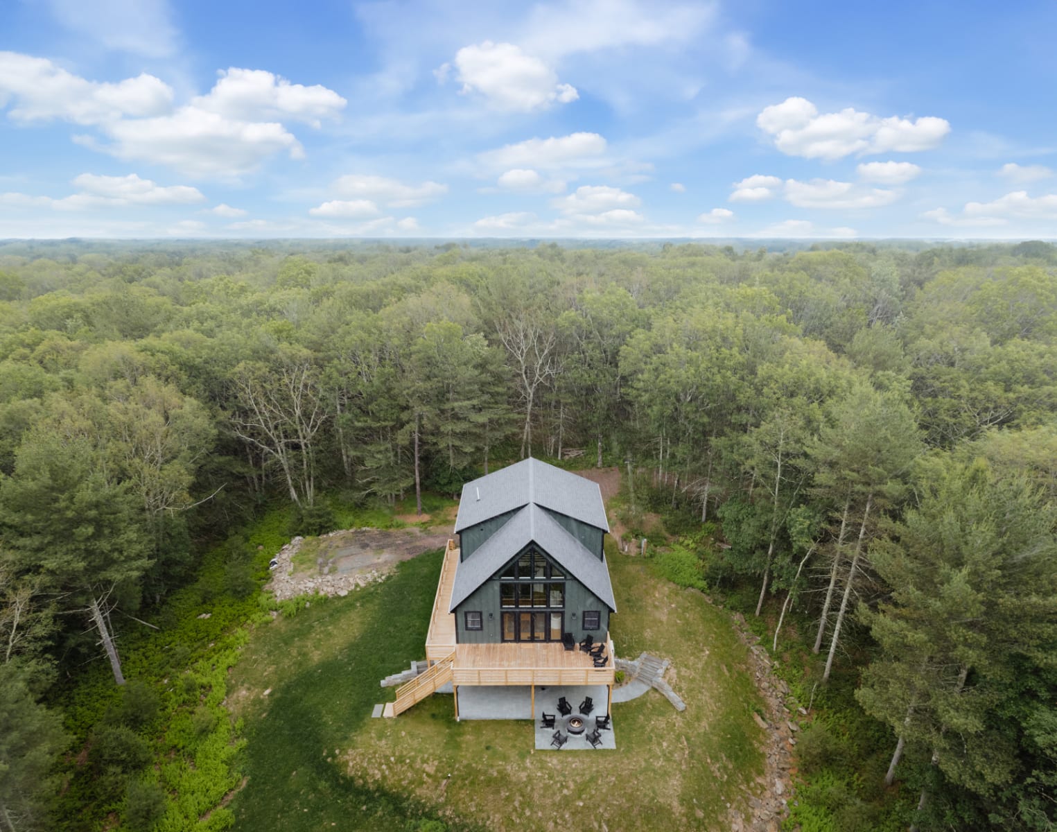 Aerial view of the modern chalet-style house with a dark roof and siding, nestled amidst a vast expanse of dense green forest under a partly cloudy sky, showcasing its secluded location.