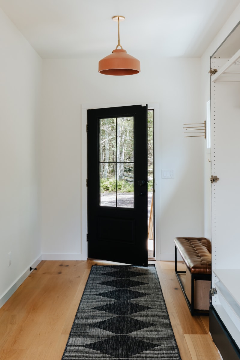 A bright foyer features light hardwood floors, white walls, and a black paneled door opening to a wooded view, complemented by a geometric runner and a tufted leather bench.