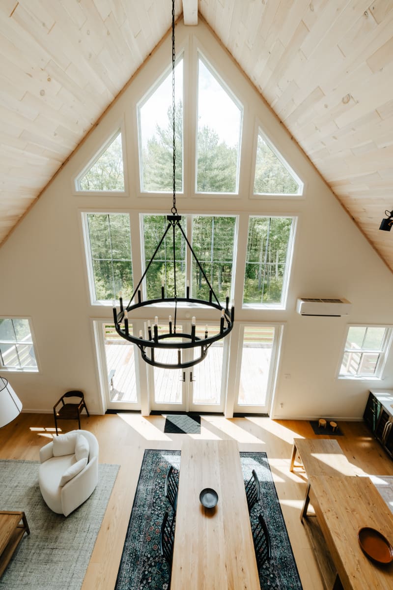An elevated view showcases the grand living and dining space with a soaring vaulted wood-paneled ceiling, a large black chandelier, and floor-to-ceiling windows offering extensive forest views.