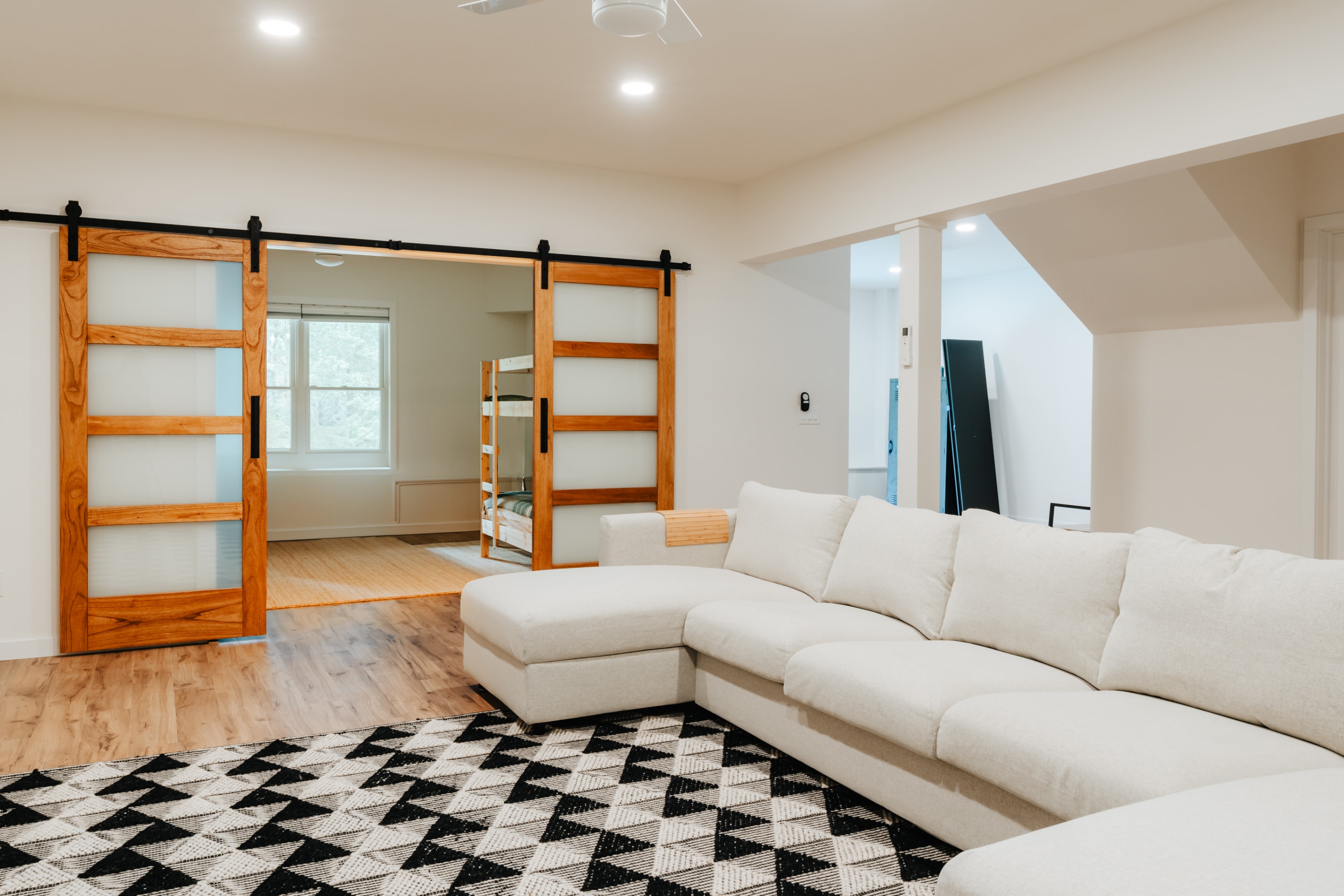 Lower-level living area with a large L-shaped white sectional sofa and a geometric area rug, featuring sliding barn doors that open to a bedroom with bunk beds, suggesting a family or game room.