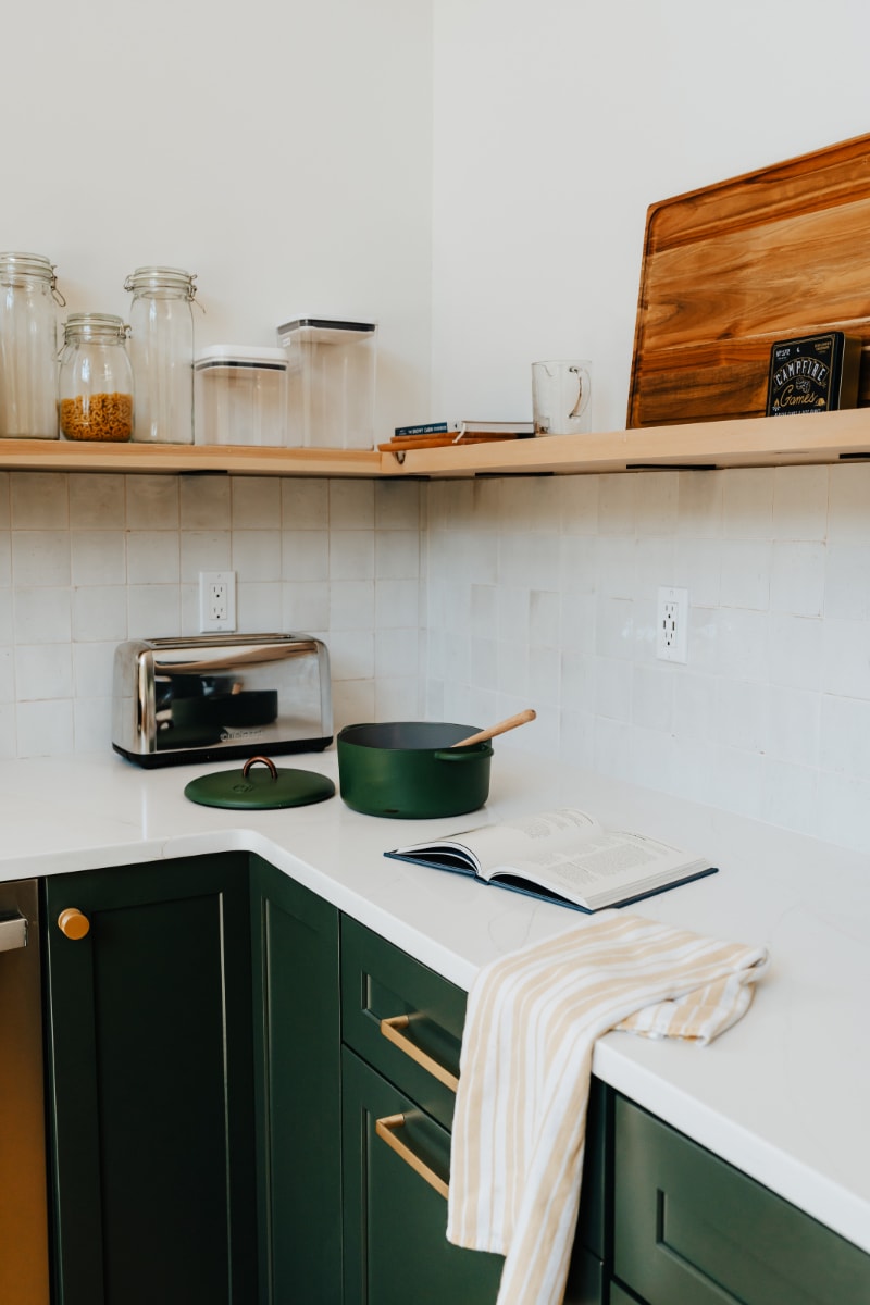 A bright kitchen corner displays white quartz countertops, dark green base cabinets with gold hardware, and open wooden shelving adorned with glass jars and kitchen essentials.