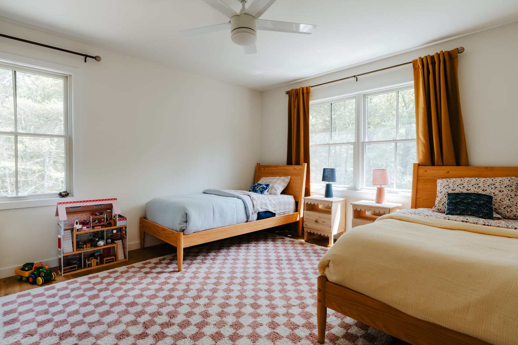A wider view of the children's bedroom, featuring two wooden twin beds, a large window with orange curtains, a pink and white checkered rug, and a dollhouse with toys in the corner, all under a white ceiling fan.
