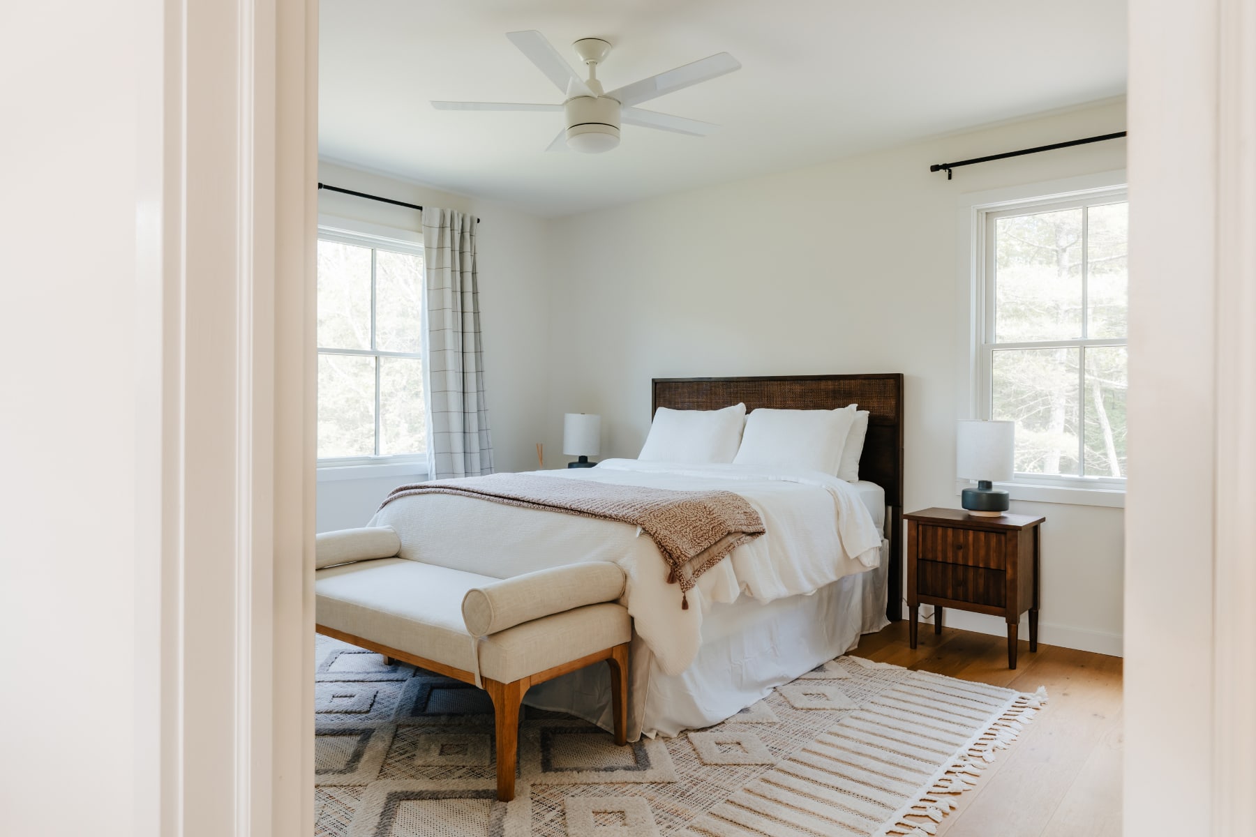 A wide shot of the bright bedroom, showcasing a dark wooden bed with white bedding and a pink throw, a light-colored upholstered bench at the foot, and a geometric patterned rug on the hardwood floor, with a ceiling fan visible.