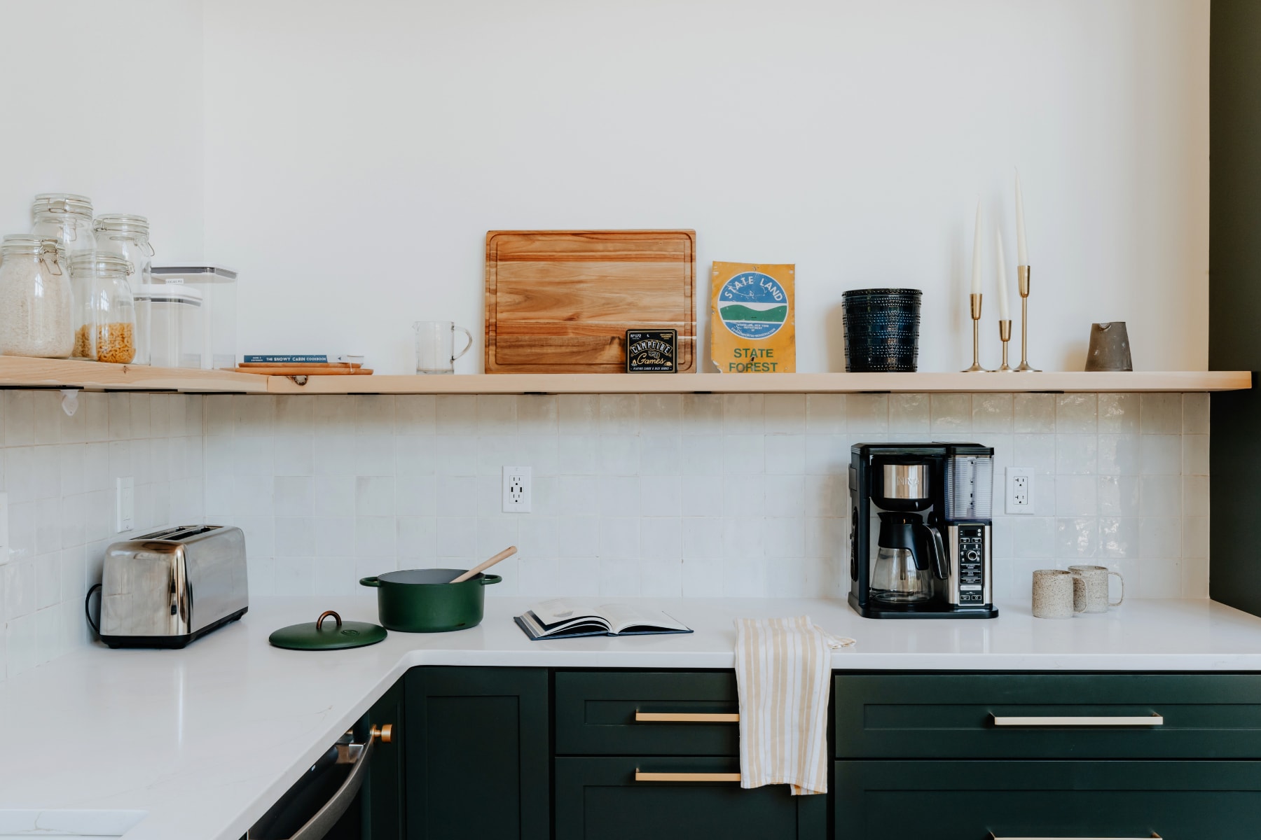 A well-appointed kitchen counter showcases white quartz surfaces, dark green cabinets, open wooden shelves with various kitchen items, and a coffee maker next to a green pot.