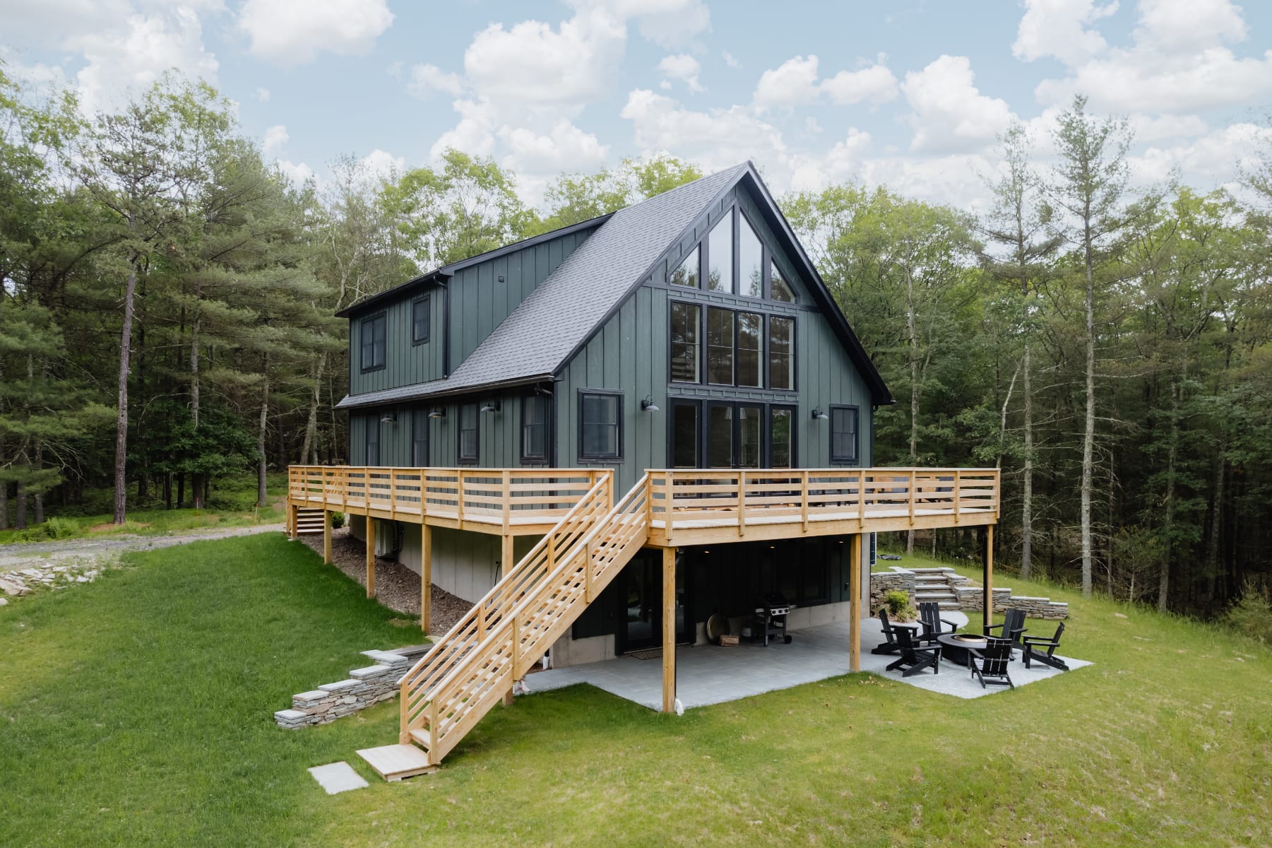 Rear view of the modern dark green chalet, featuring a large elevated wooden deck with stairs descending to a lower patio area with outdoor seating, all surrounded by a lush green lawn and forest.