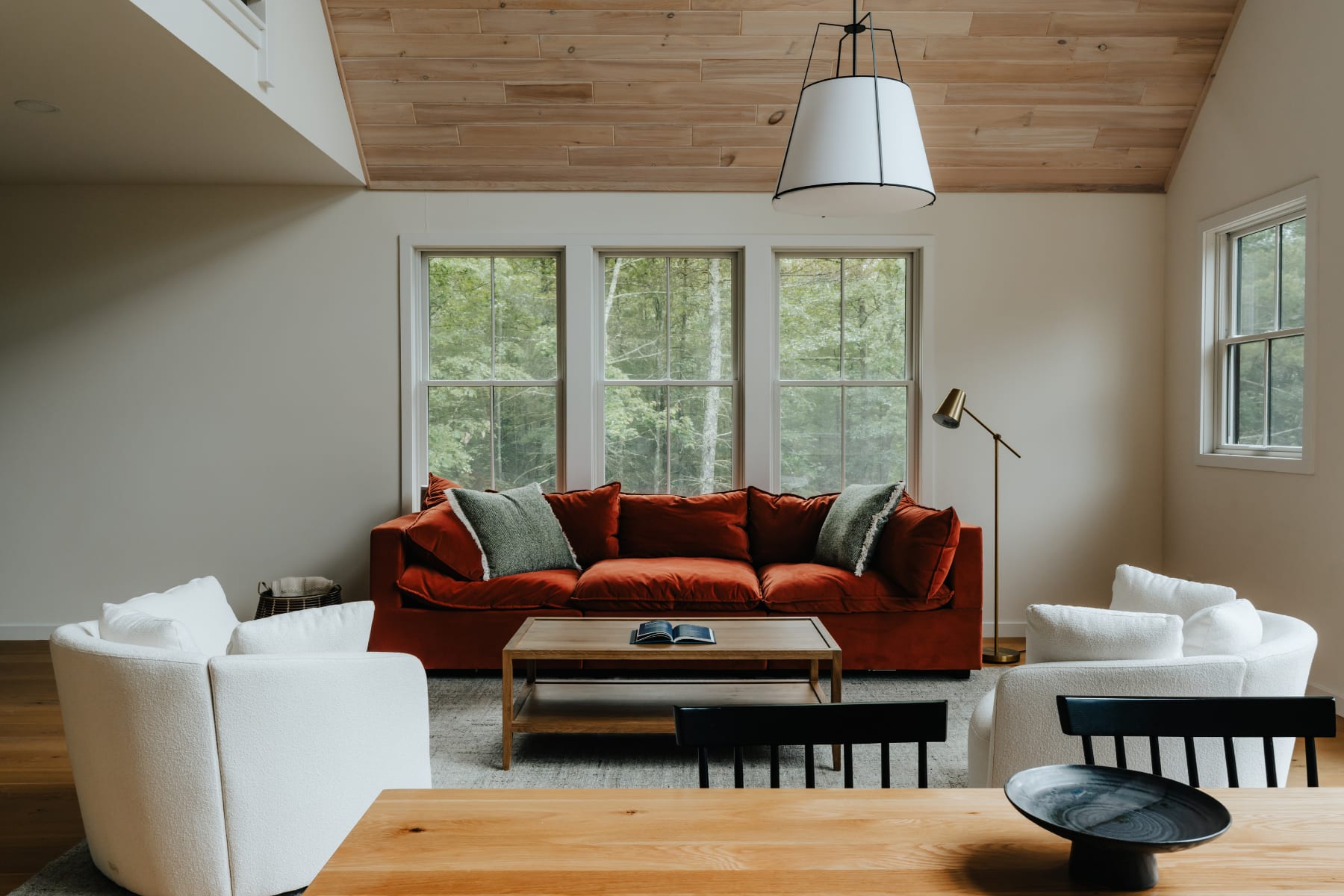 Cozy living room with a vaulted wood-plank ceiling, a large rust-colored velvet sofa, two white armchairs, a wooden coffee table, and multiple windows providing views of the surrounding trees.