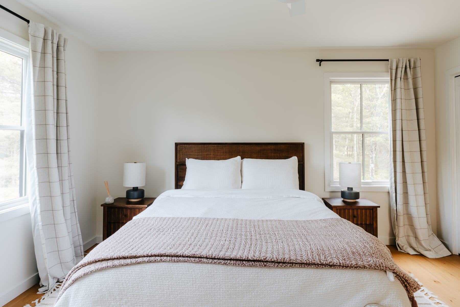 A bright and airy bedroom featuring a dark wooden headboard, crisp white bedding with a textured pink throw, flanked by two wooden nightstands with modern lamps, and two large windows with light plaid curtains.