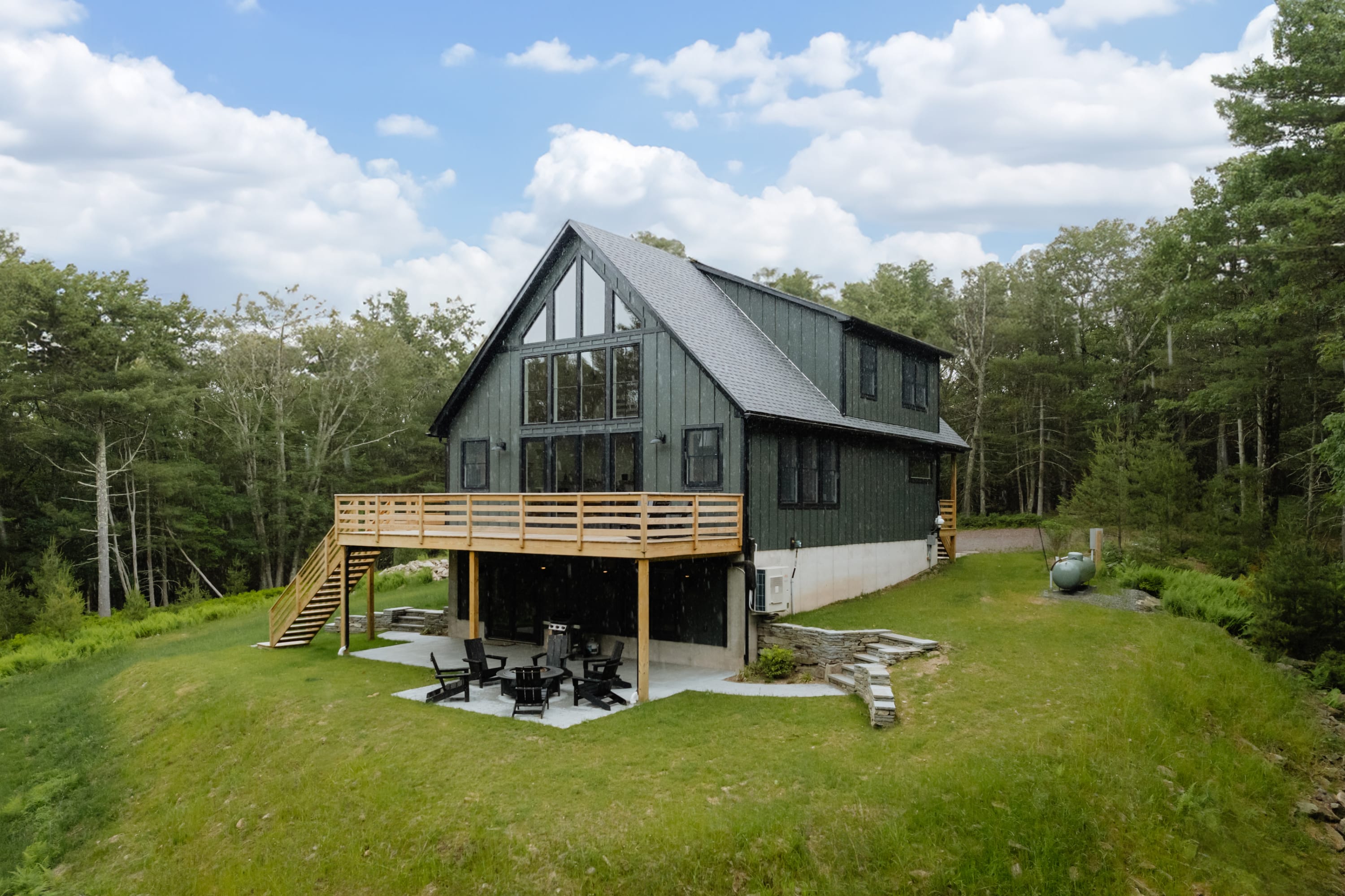 Aerial view of the modern dark green chalet, highlighting its elevated wooden deck, lower patio with seating, and expansive grassy yard, nestled within a dense forest landscape under a cloudy sky.