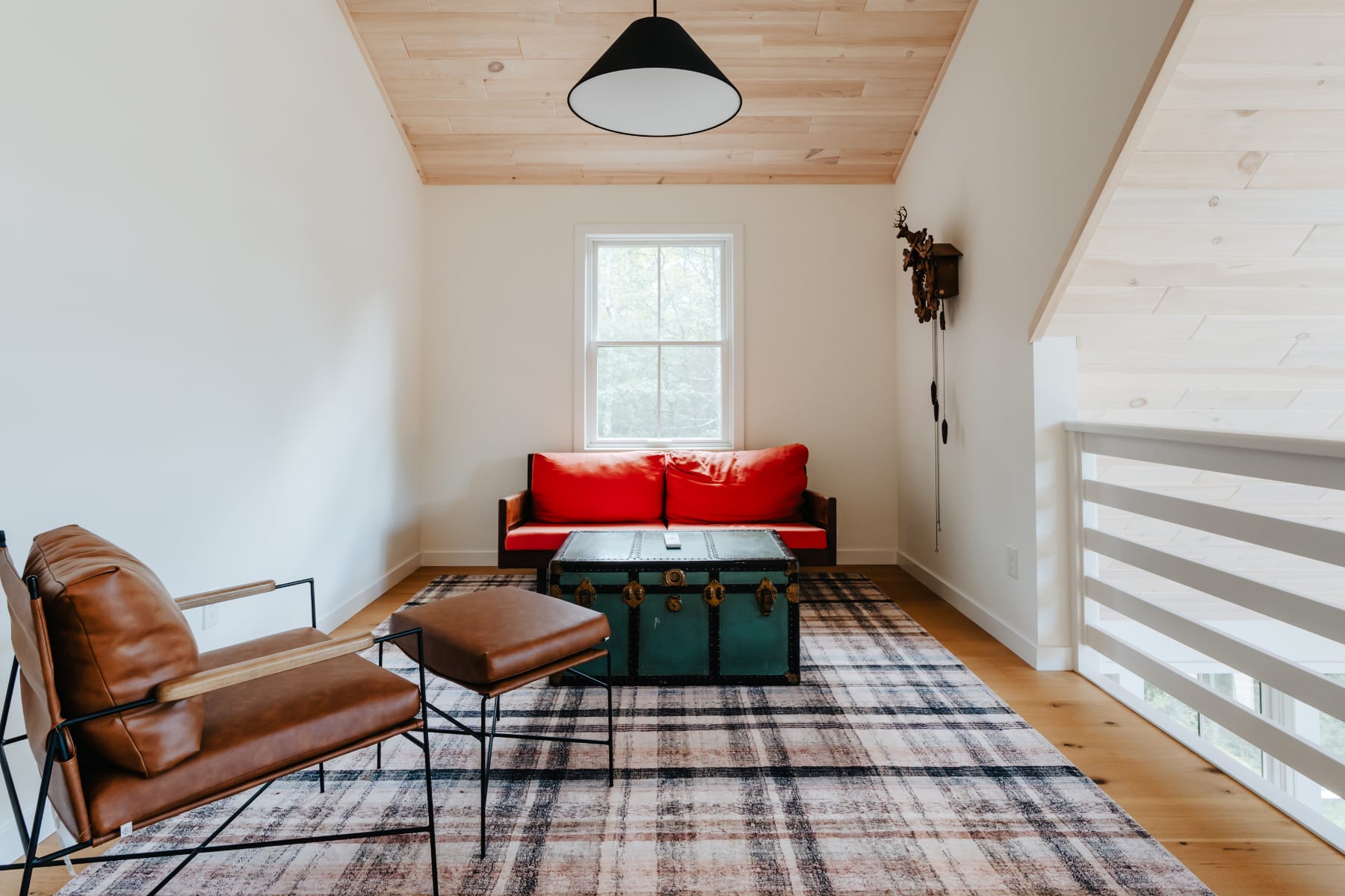 A cozy loft living area with a red sofa, a brown leather armchair, and a green vintage trunk serving as a coffee table, all set on a plaid rug under a light wood paneled ceiling. A window provides natural light, and a cuckoo clock adorns the wall.