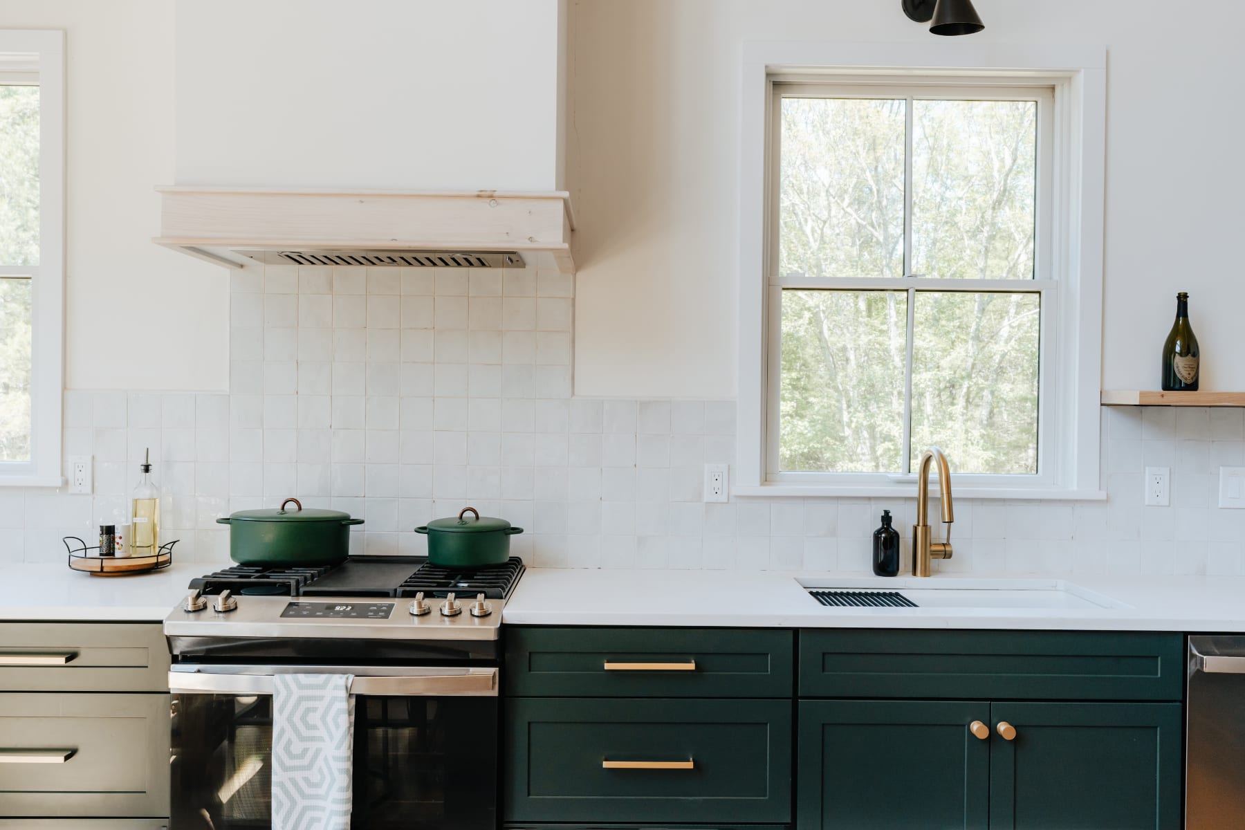 The kitchen features a stainless steel gas range with a custom wood range hood, dark green cabinetry, white quartz countertops, and a gold faucet over a white sink, all against a white tiled backsplash.