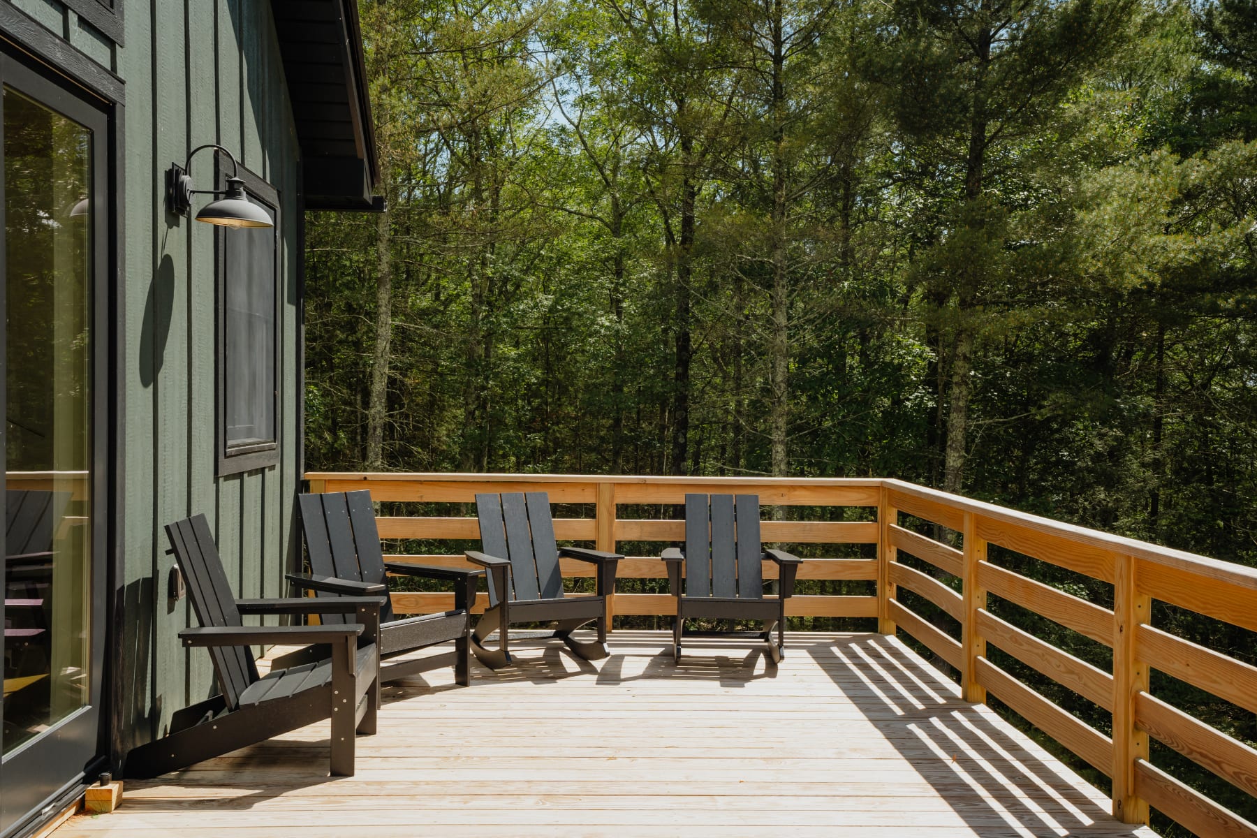 Wooden outdoor deck with a natural wood railing, furnished with three black Adirondack chairs, offering a serene view of the surrounding lush green forest.
