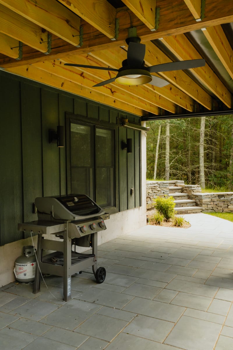 Covered outdoor patio area with a gas BBQ grill, concrete pavers, and a wooden ceiling with exposed beams and a ceiling fan, overlooking a wooded area.