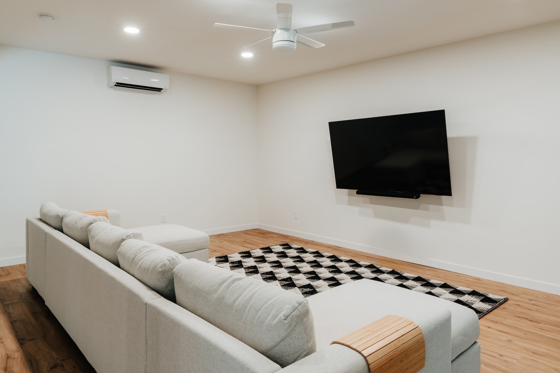 A modern media room featuring a large wall-mounted television with a soundbar, a comfortable light grey sectional sofa, and a black and white geometric rug on the hardwood floor, with a ceiling fan and a mini-split AC unit.