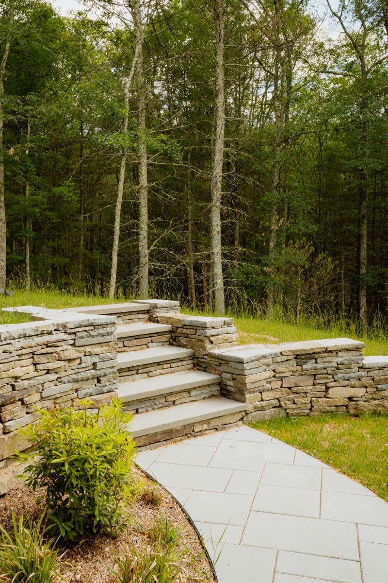 Landscaped outdoor area featuring a winding stone paver pathway leading to multi-tiered stone steps and retaining walls, set against a backdrop of lush green grass and dense forest.
