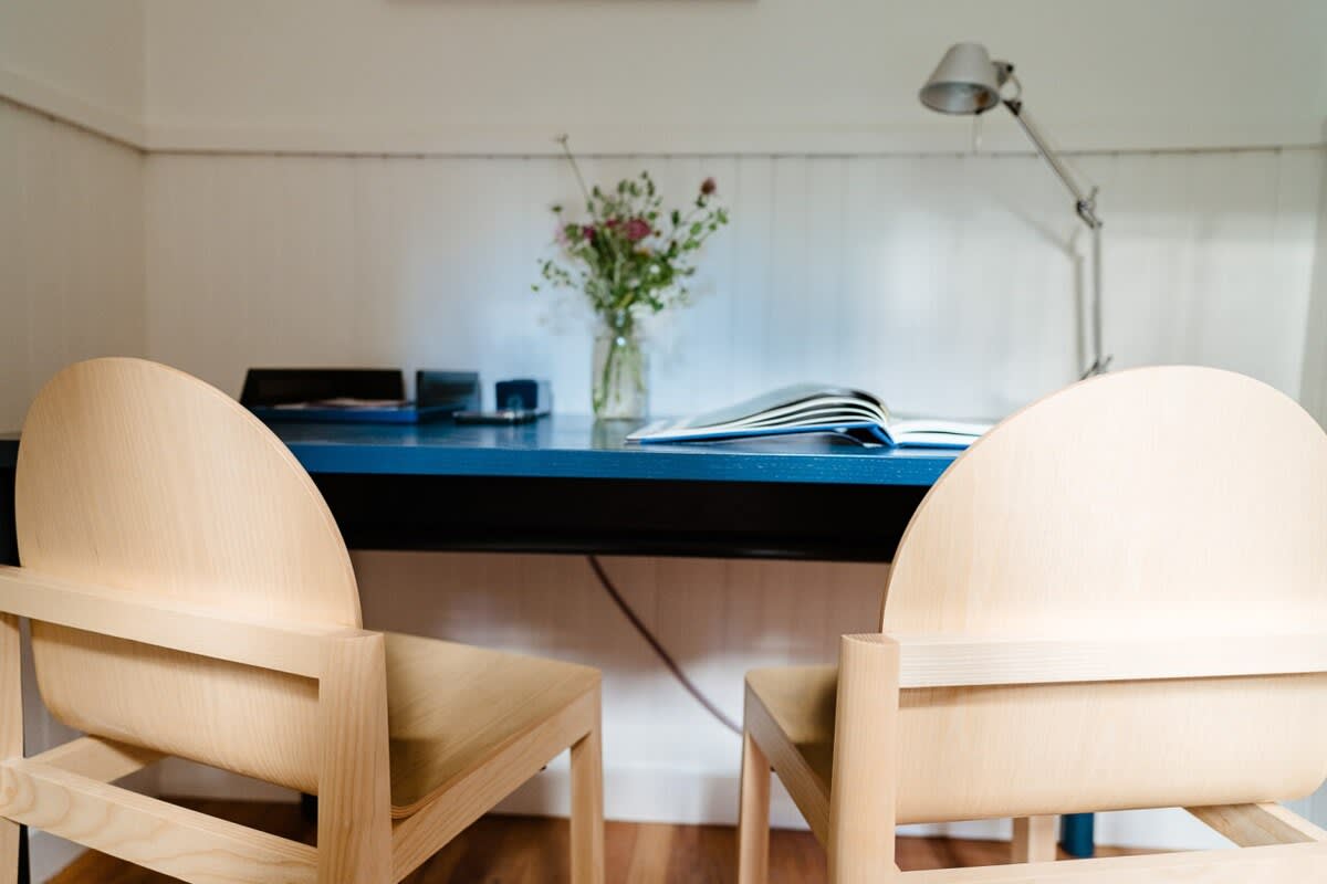 A dedicated workspace features a vibrant blue desk with two light wood chairs, a desk lamp, and a vase of flowers. The room has white paneled walls and hardwood floors, creating a bright and functional area.